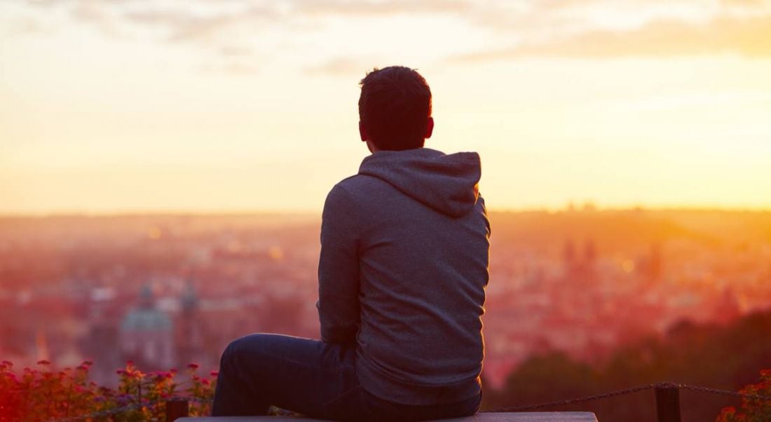 A young man wearing a hoodie. His back is to camera and he is looking at a sunrise and thinking about his life experiences.