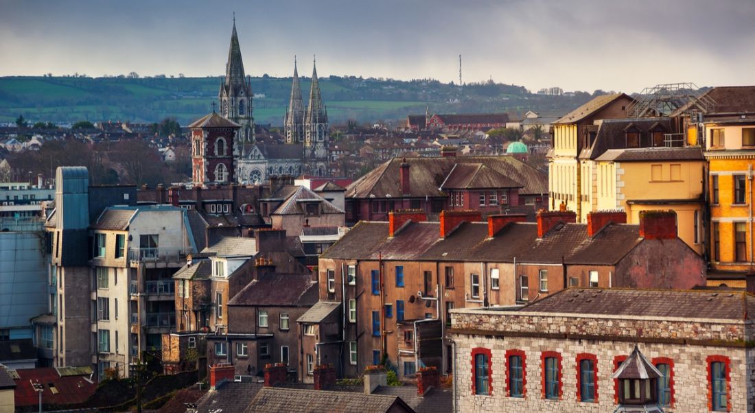 A vantage point view of an old part of Cork city, with a church and rolling emerald hills in the background.