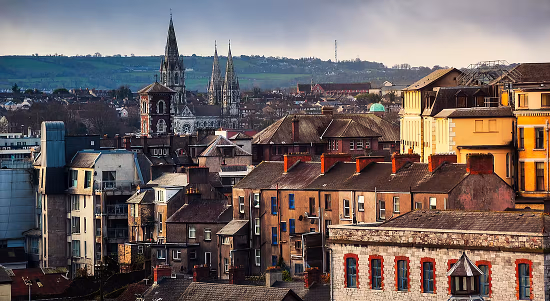 A vantage point view of an old part of Cork city, with a church and rolling emerald hills in the background.