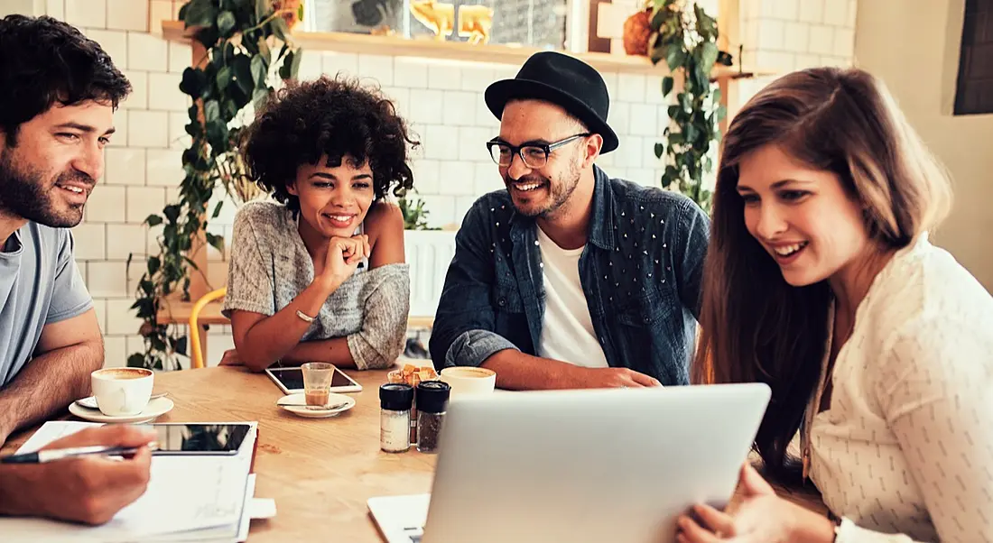 Group of friends hanging out in chic coffee shop huddled around a slim, silver laptop.