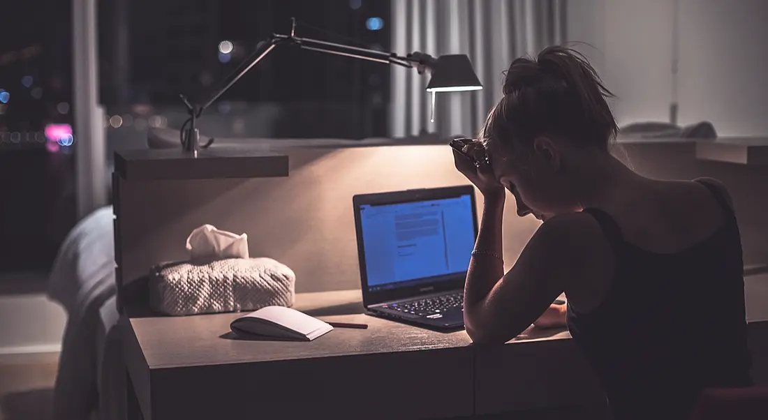 A young woman in a tank top sitting at her desk in front of her laptop with her hand on her forehead, looking worn out.