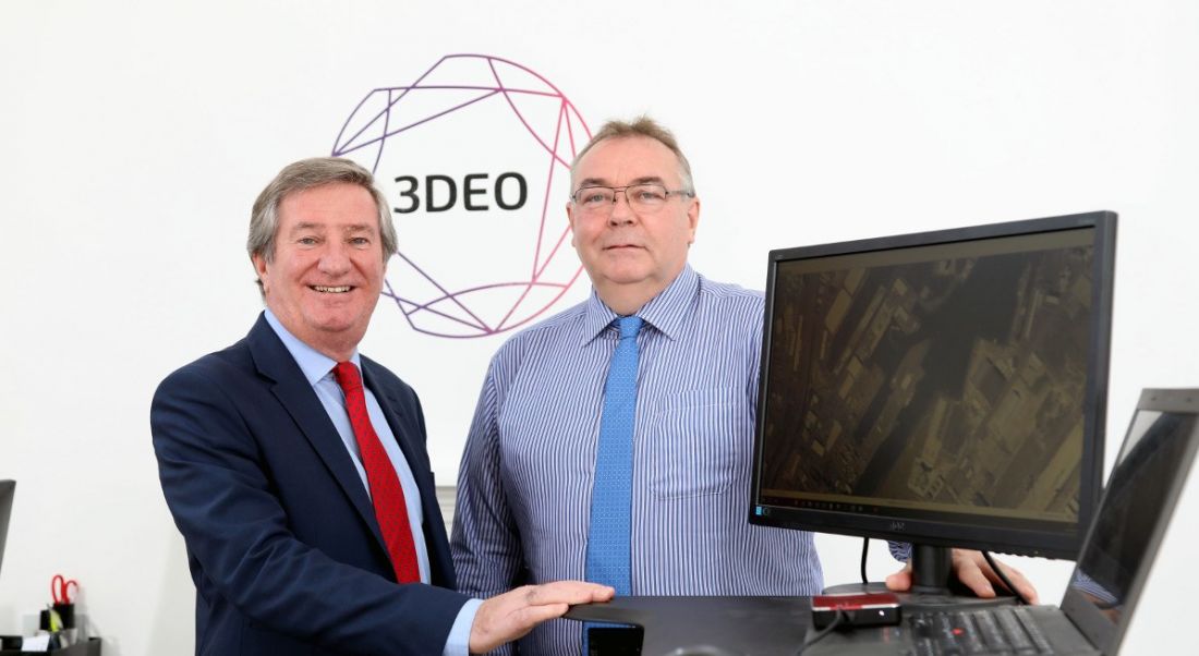 Two middle aged men in shirts and dies standing next to a desktop in front of a geometric logo with ‘3DEO’ inside it.