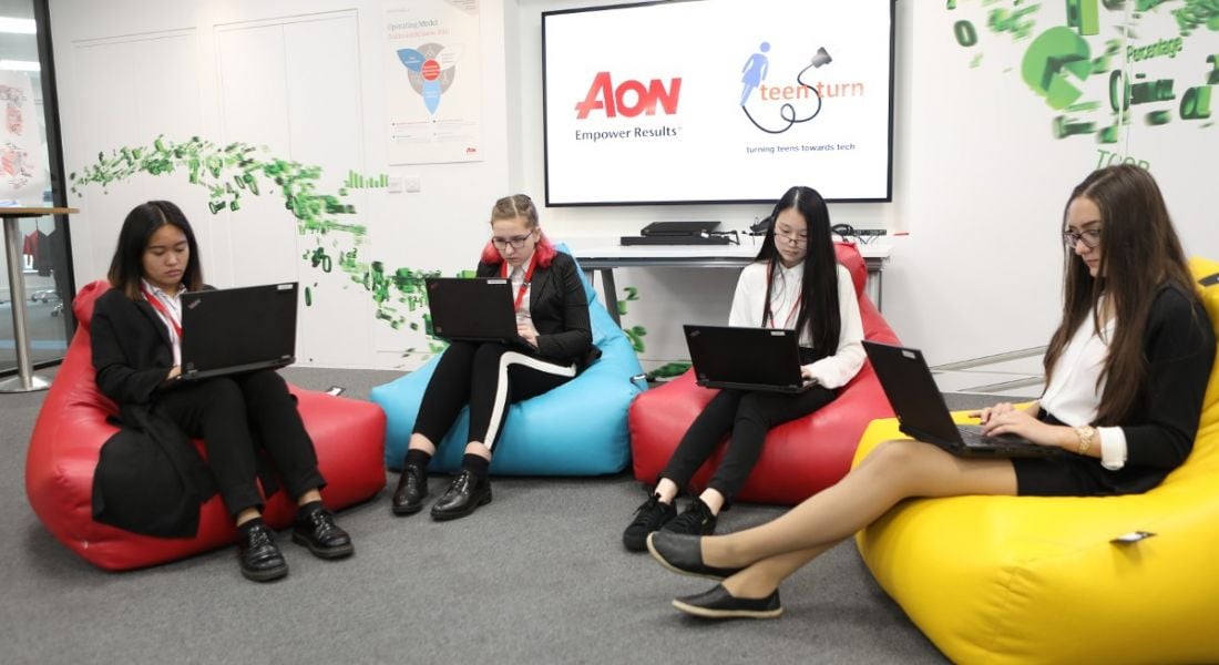 Four young women sit on bean bags while looking intently at their laptops.