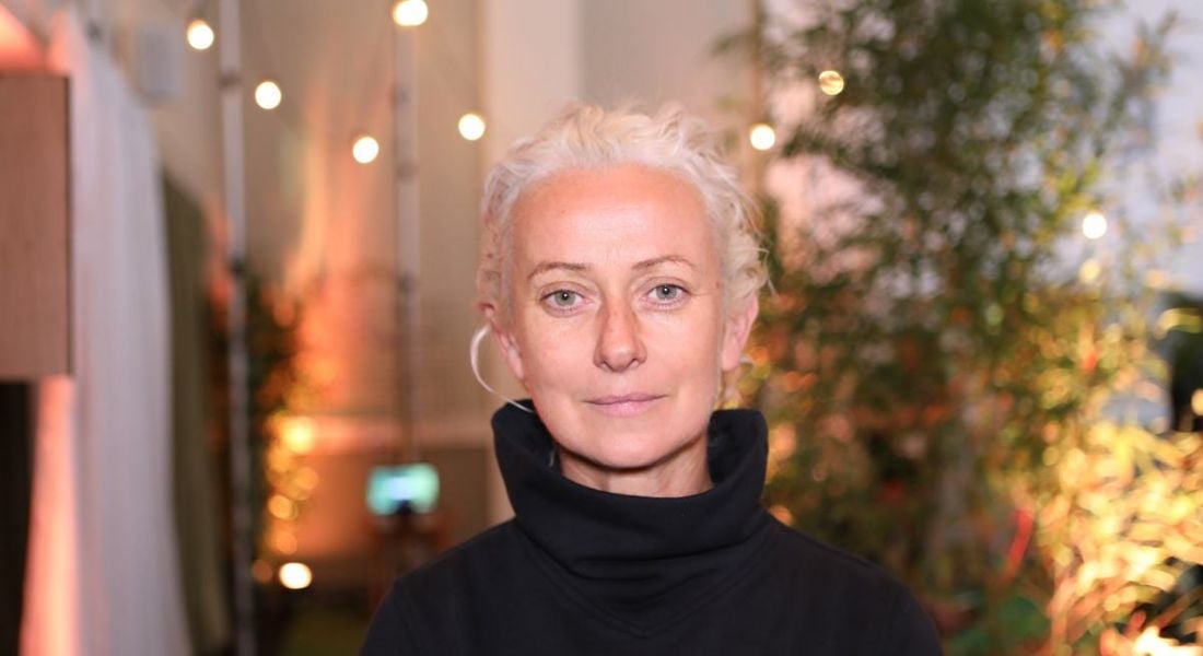 Headshot of a woman with white hair pulled back off her face against a background of plants and fairy lights inside a grand room.