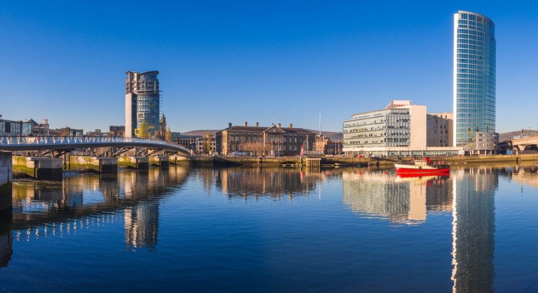 Belfast cityscape with a bridge to the left, buildings in the centre and right, and clear still water in the foreground and a small red and white boat on the water. There is a bright blue sky in the background.