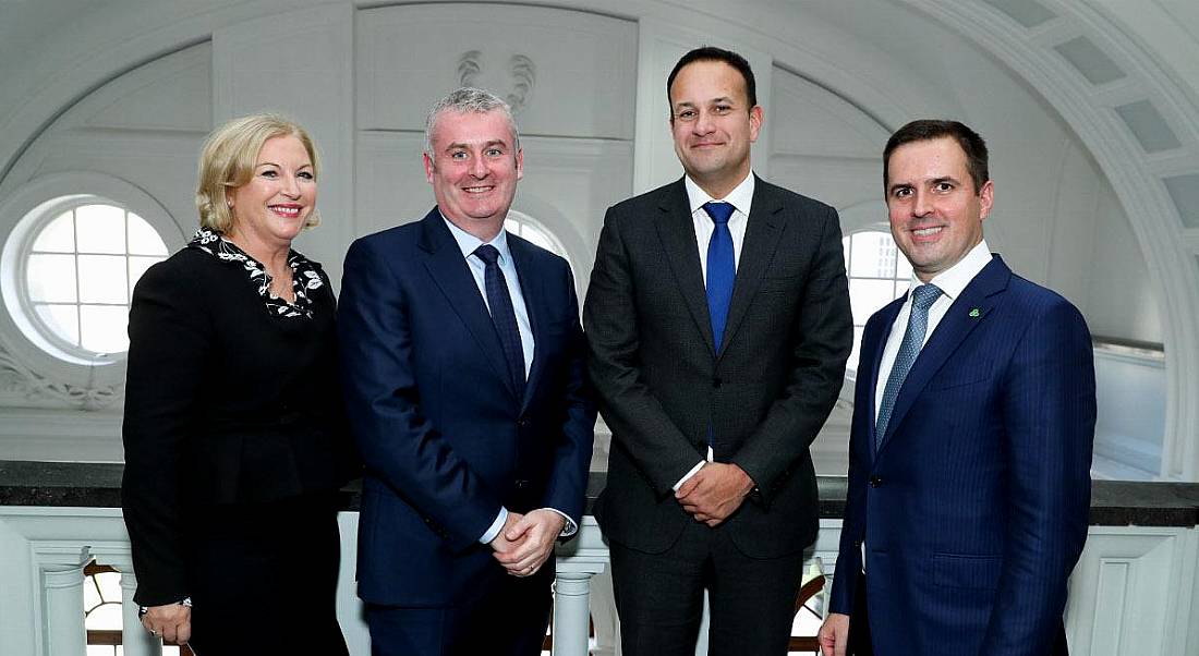 A blonde woman in a black skirt and blazer and three men in suits standing in a line smiling at the MSD jobs announcement.