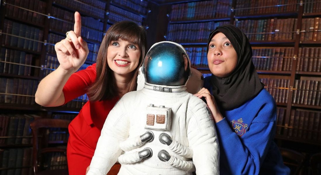 Attractive woman in red dress standing with an astronaut suit beside a 13-year-old girl. Both are looking up in awe.