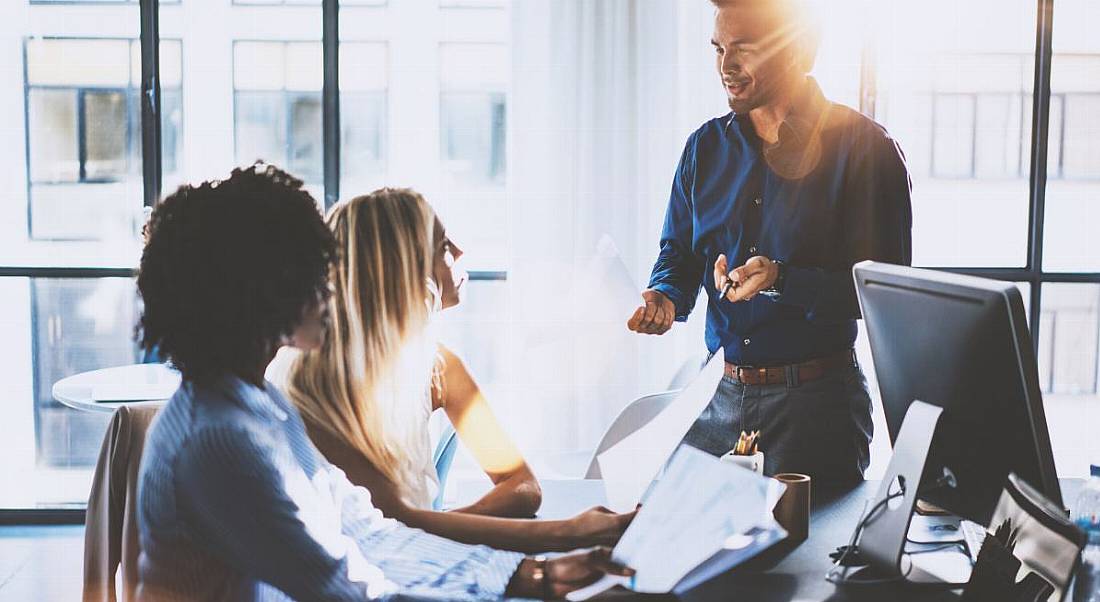 A group of young professionals working at a large enterprise holding papers and chatting at their desks about work.