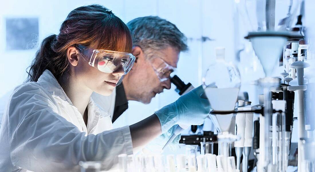 A brunette scientist wearing protective goggles hunched over test tubes, an example of Women in STEM.