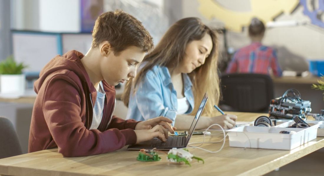 A young boy coding on a laptop next sitting next to a young girl writing in a classroom setting.