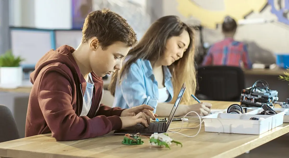 A young boy coding on a laptop next sitting next to a young girl writing in a classroom setting.