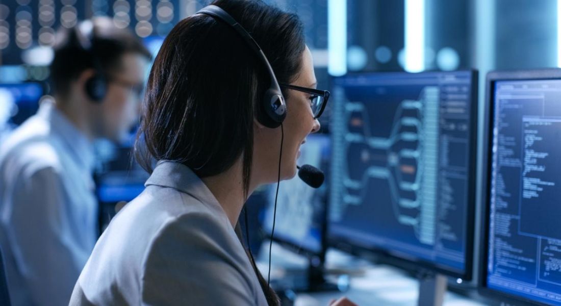 The back of the head of a dark-haired woman with glasses wearing a head set and looking at a computer screen while working in a technical support role.