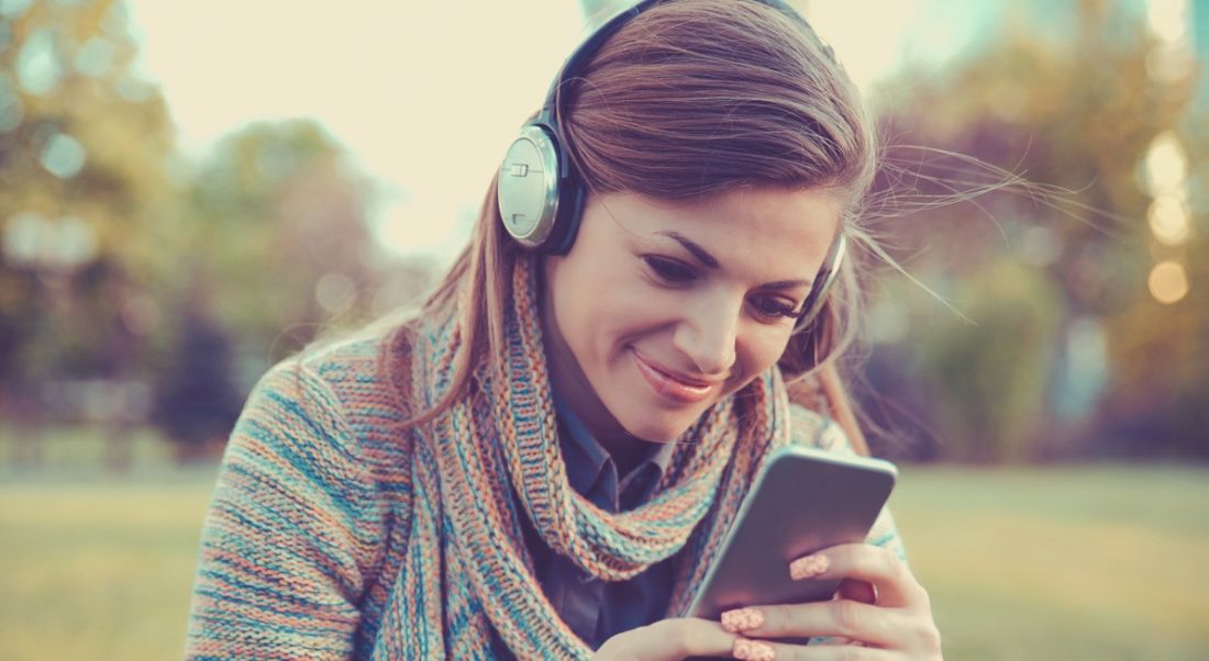 A smiling young woman sitting in a park looking at a mobile with headphones on, depicting gamification.