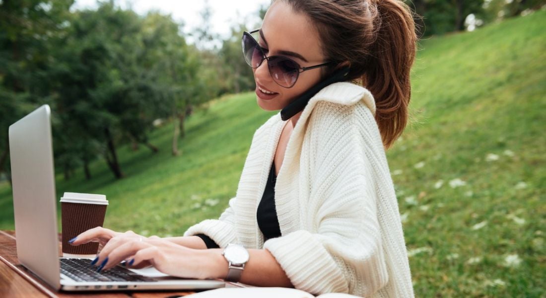 A young woman wearing sunglasses typing on her laptop with her smartphone pressed between her ear and shoulder.