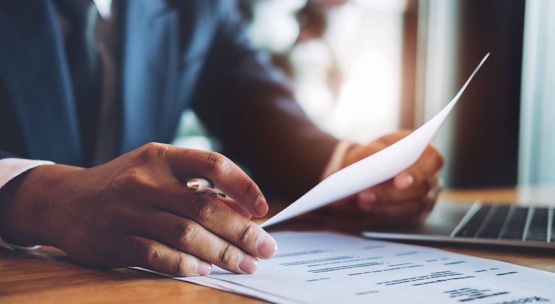 Close-up shot of a suited man’s hands as he looks at a piece of paper – his CV – with a laptop beside him.