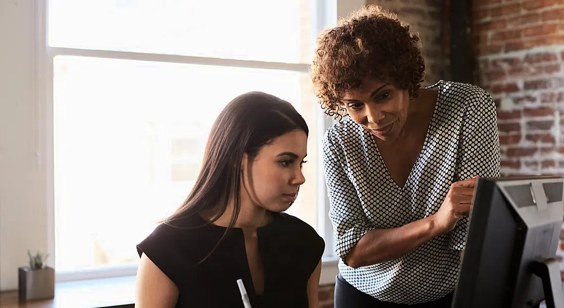 One woman acting as a mentor standing beside another woman sitting at a computer.