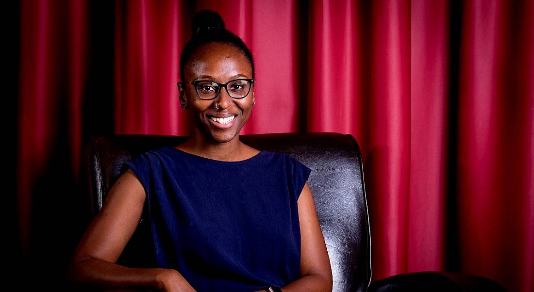 A smiling young black woman with her hair up, wearing a blue top, sitting in a leather chair with a red curtain behind her.