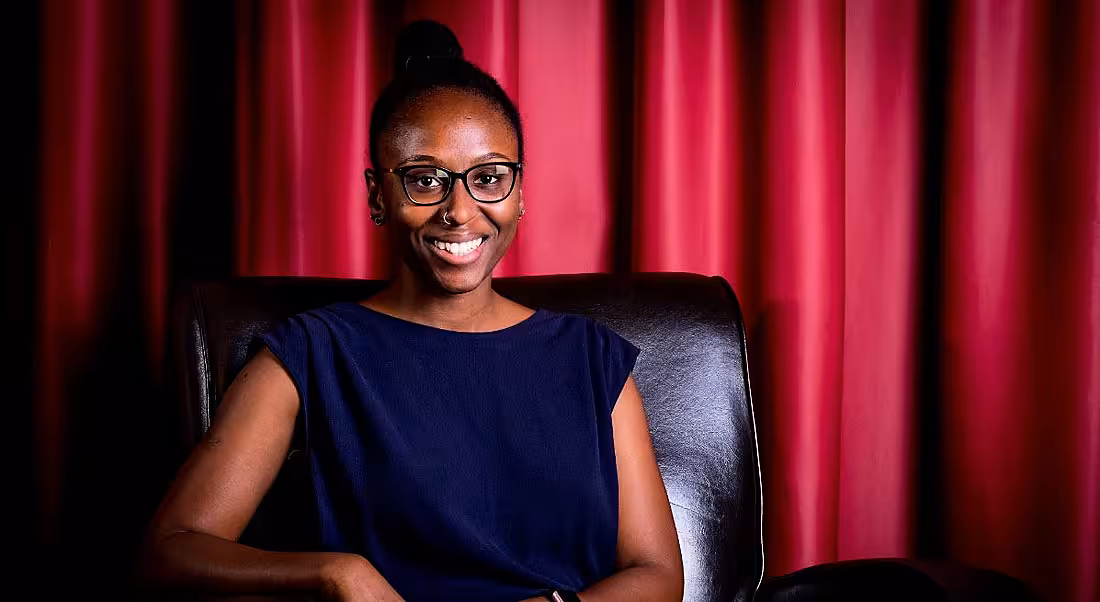 A smiling young black woman with her hair up, wearing a blue top, sitting in a leather chair with a red curtain behind her.