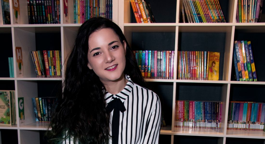 A dark-haired young woman from the gaming industry smiling at the camera against a backdrop of shelves filled with books.