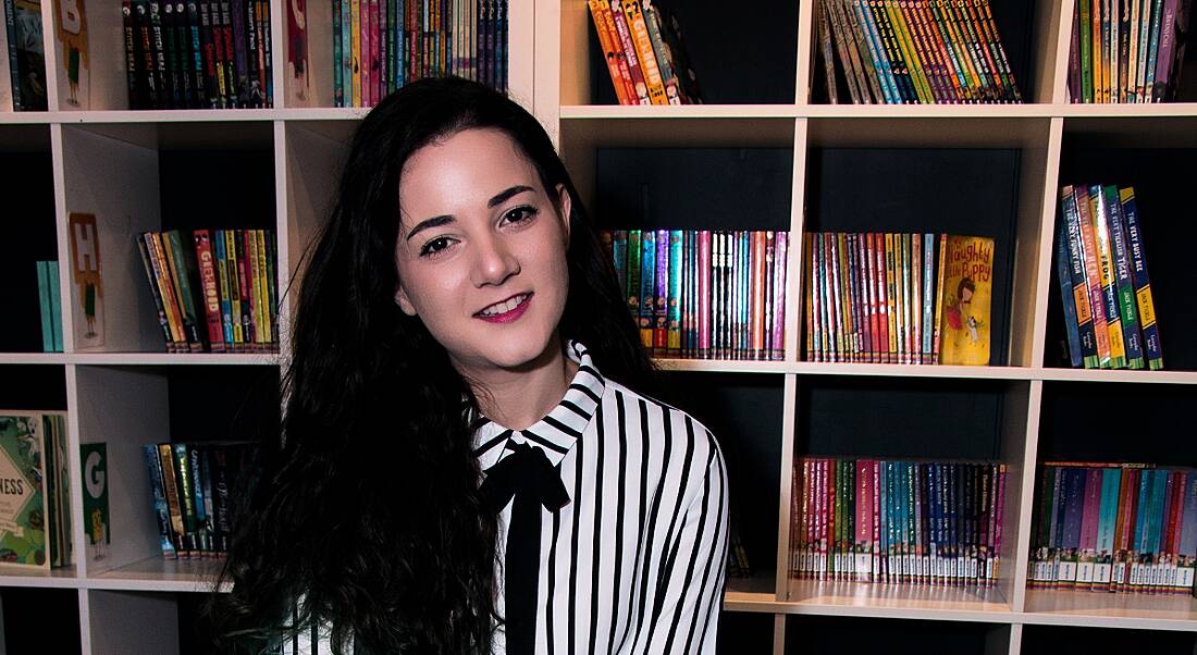 A dark-haired young woman from the gaming industry smiling at the camera against a backdrop of shelves filled with books.