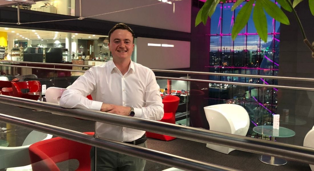 Smiling young man in a white shirt, leaning on a metal barrier in a bright PwC office surrounded by colourful chairs.