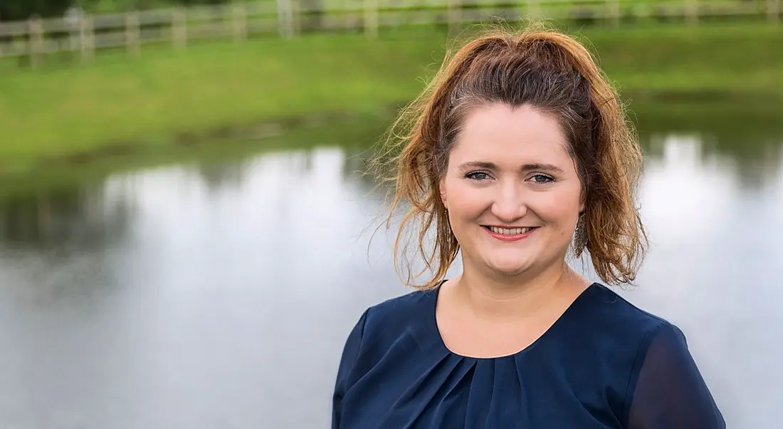 Headshot of a smiling woman from Pramerica with red hair tied up against a background of grass and a lake.