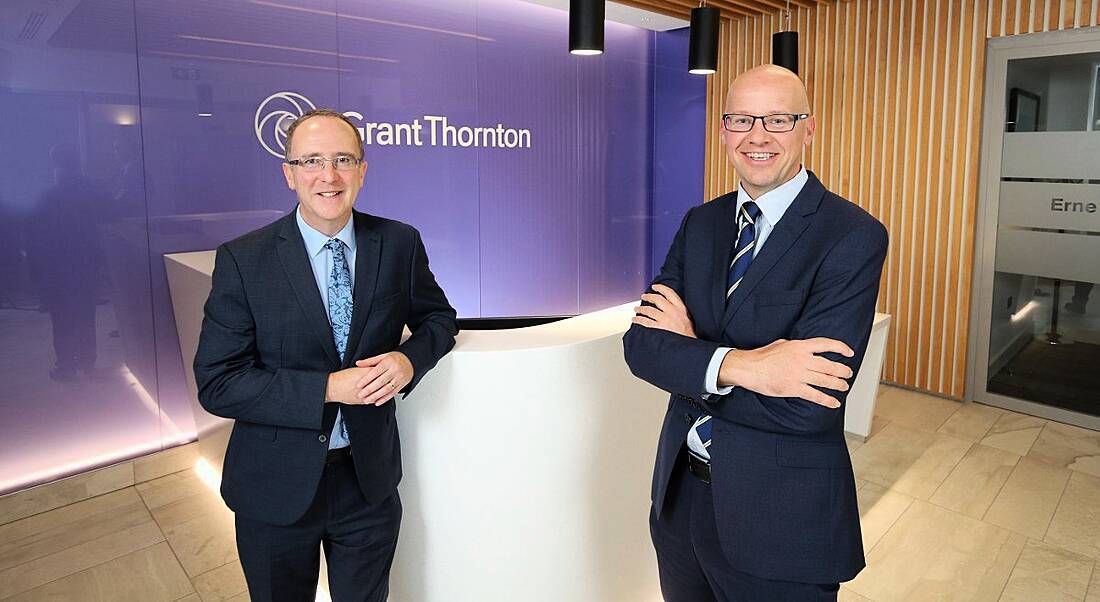 Two suited glasses wearing men smiling at the camera against a purple wall emblazoned with company name Grant Thornton.