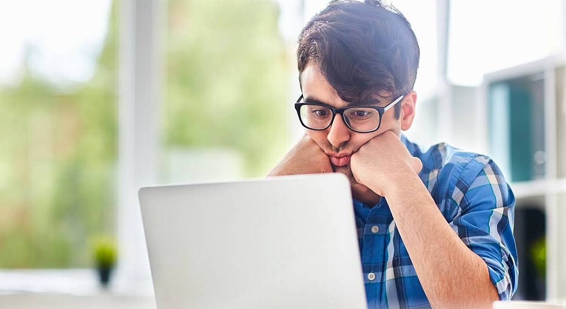 A young man dressed casually, looking very deflated at a laptop searching for an entry-level job with his head on his hands.