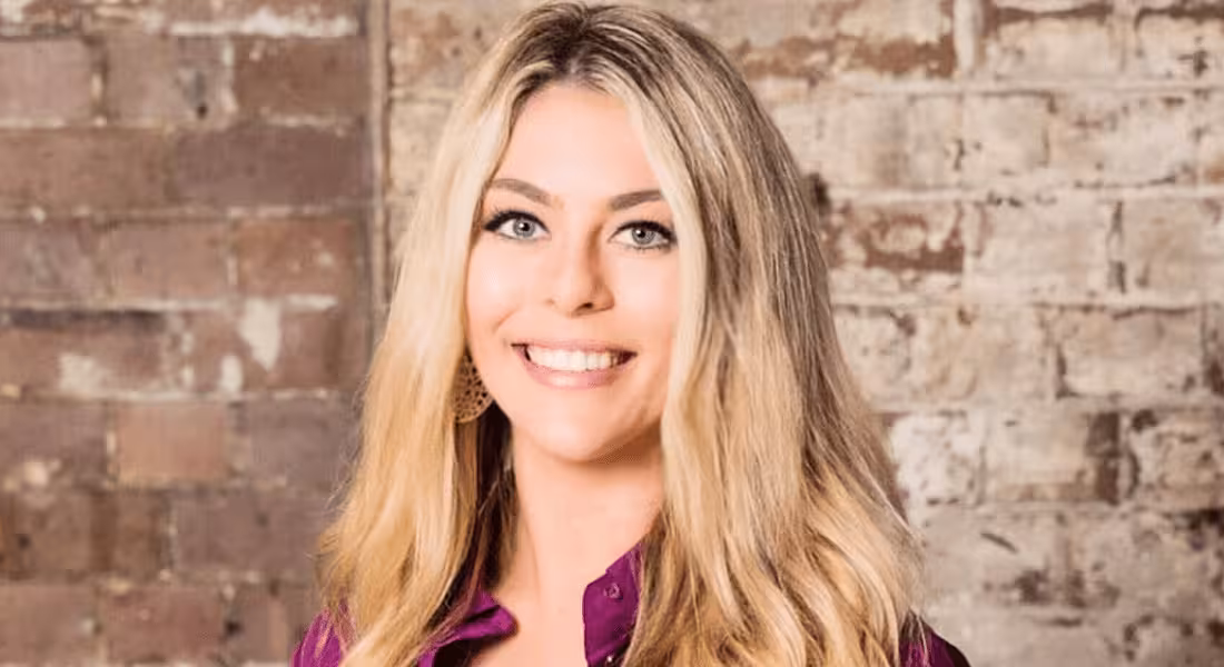 A headshot of a smiling young woman with long blonde hair against a blurred-out brick wall background.