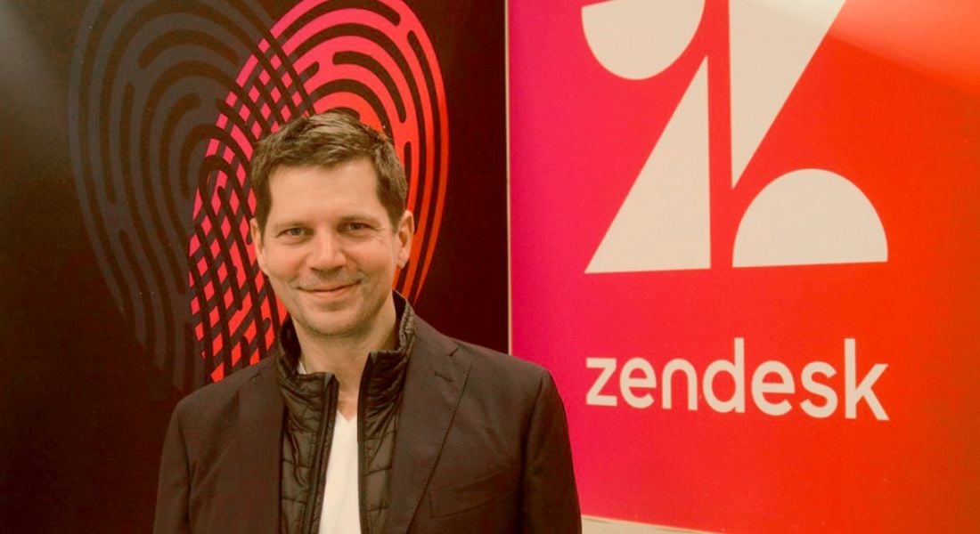 A young man with brown hair smirking into the camera with a red wall emblazoned with the Zendesk logo behind him.
