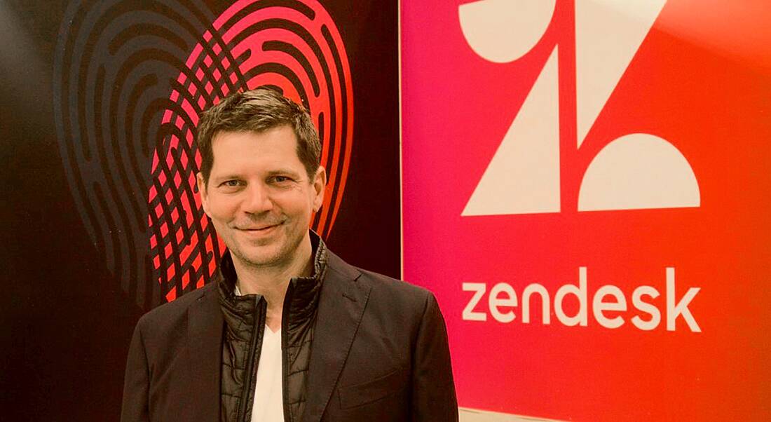 A young man with brown hair smirking into the camera with a red wall emblazoned with the Zendesk logo behind him.