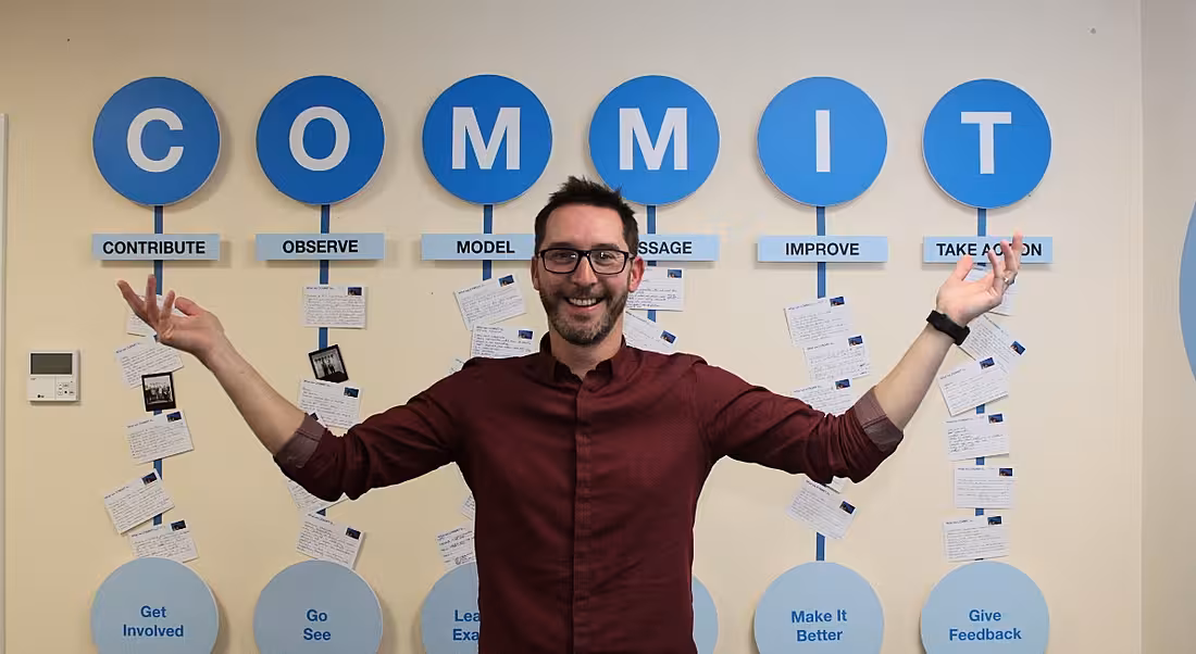 A young bearded man with thick-rimmed glasses with arms aloft with the word ‘Commit’ emblazoned on the wall behind.