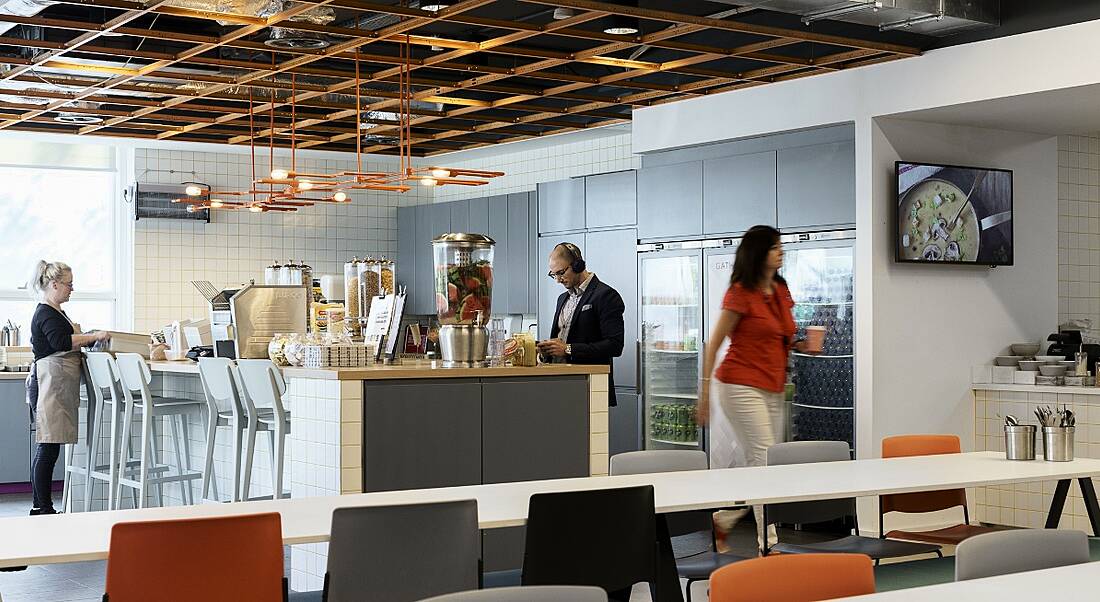 A view of an spacious well-lit modrn office kitchen buzzing with people gathered around a water cooler teaming with ice, water and fresh fruit.