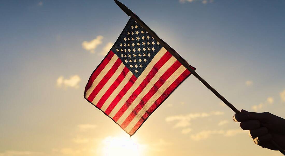 A hand holding a small US flag against the backdrop of a rising sun.