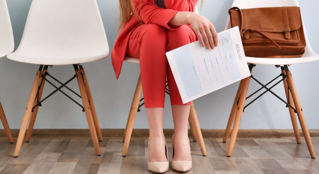 View of a woman’s legs and a hand cliutching as résumé as she sits in a waiting room before a job interview.