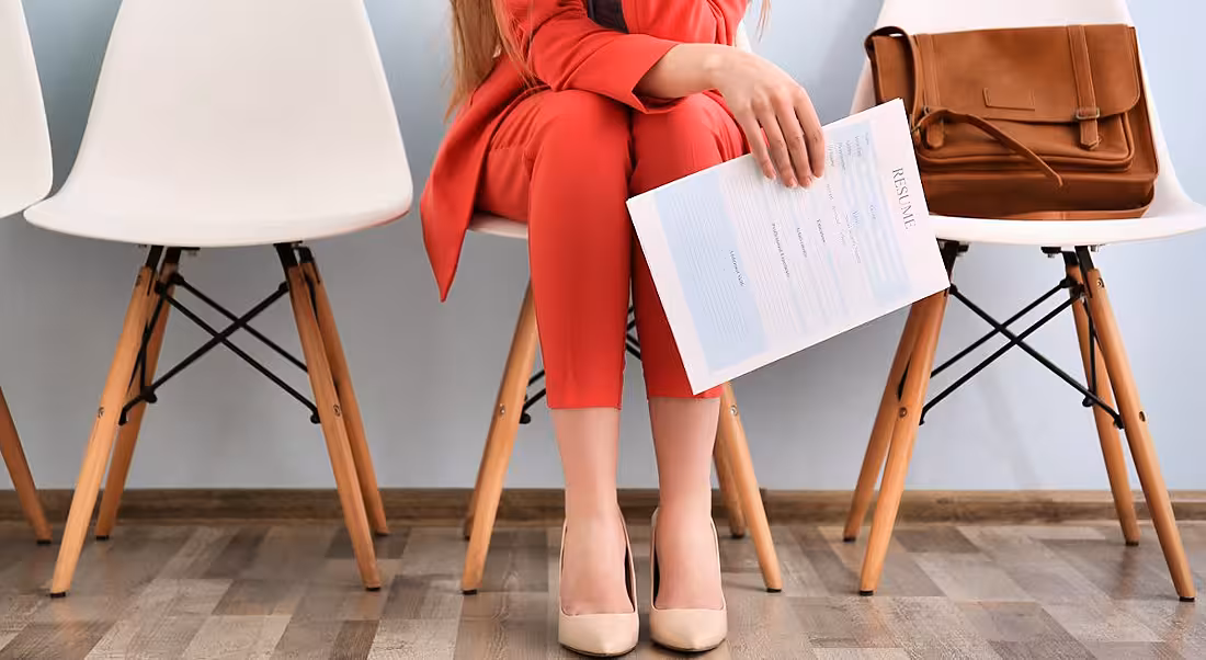 View of a woman’s legs and a hand cliutching as résumé as she sits in a waiting room before a job interview.