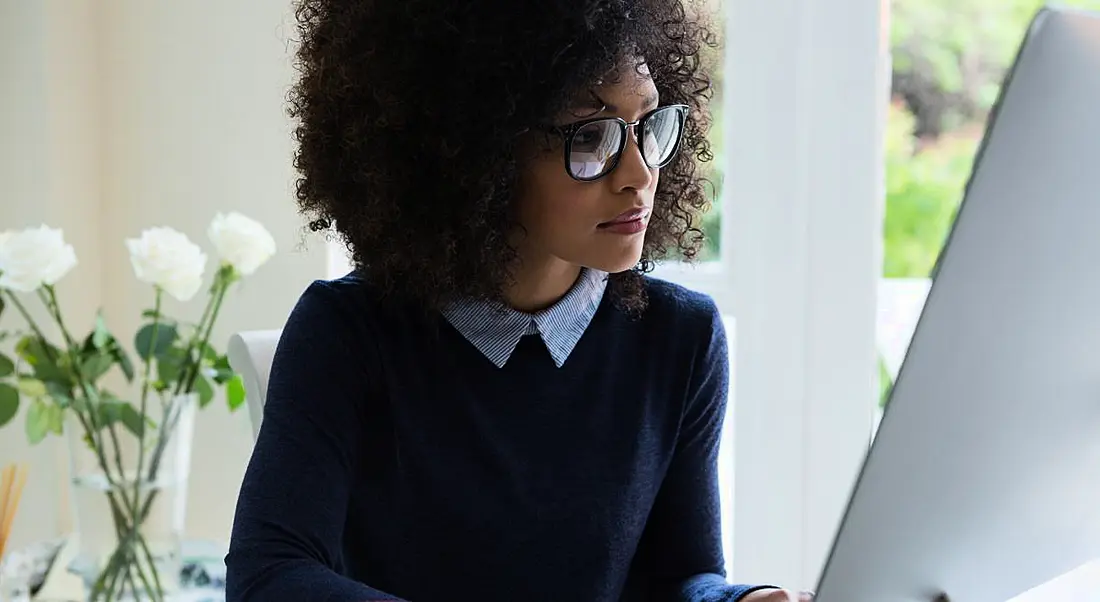 A young woman with glasses sitting at her desk working with a vase of white roses behind her.