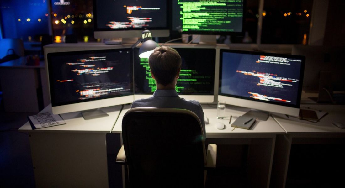 The back of a young blonde man’s head as he stares at a number of computer monitors illuminated with code. He works in cybersecurity.