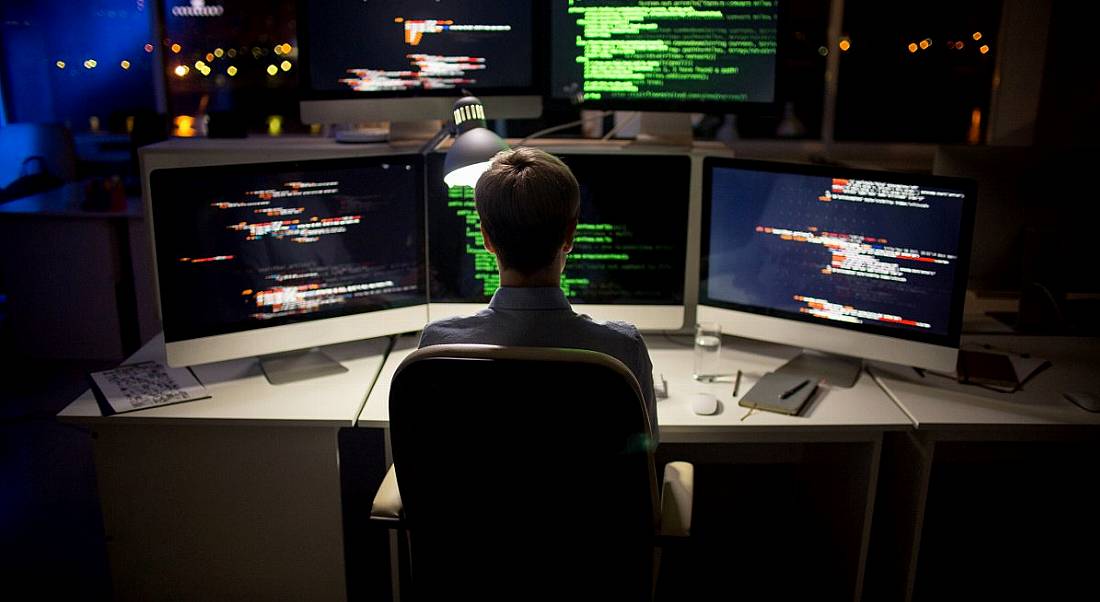 The back of a young blonde man’s head as he stares at a number of computer monitors illuminated with code. He works in cybersecurity.