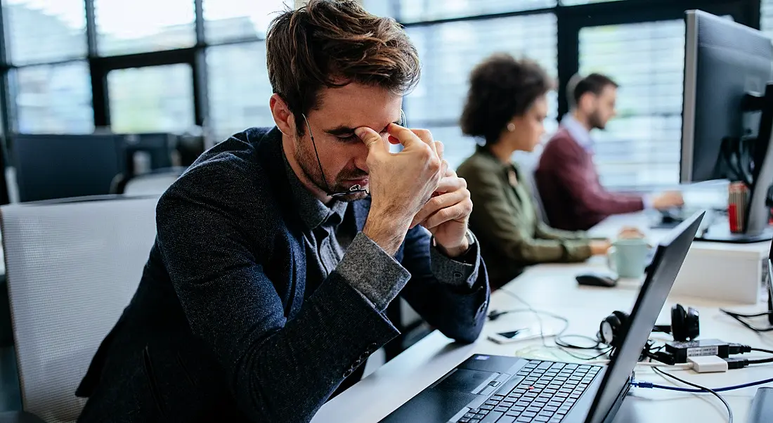 A frustrated man sitting in an office in front of a laptop pinching the bridge of his nose. He has been in his job too long.