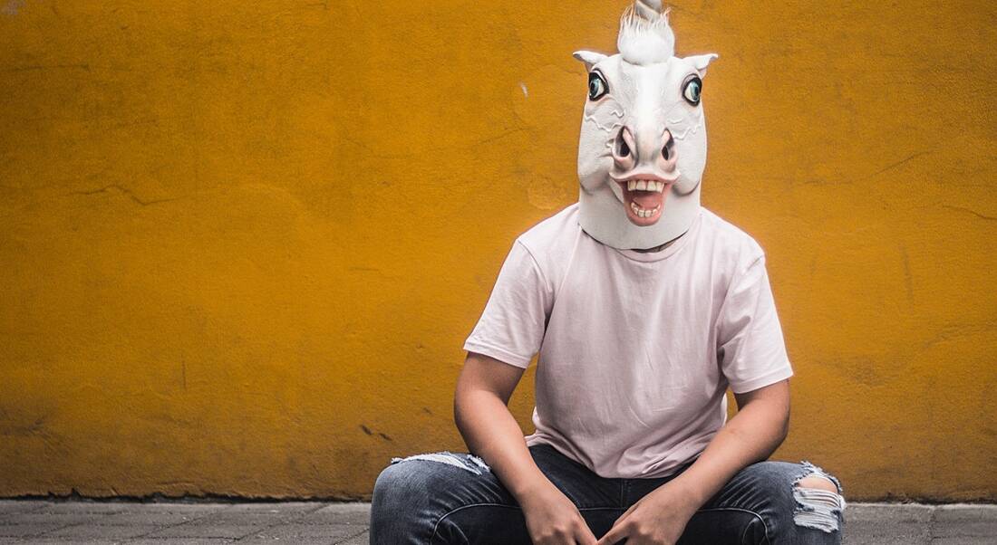 Man in unicorn mask sitting against yellow wall.