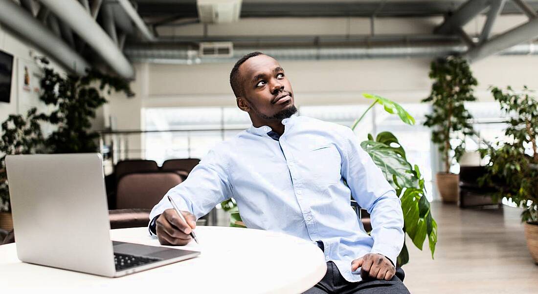 A man in a white shirt leaning back in his chair looking pensively into the difference while working on his laptop.