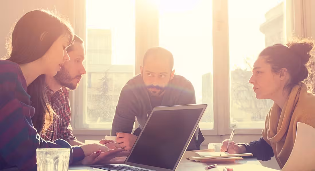 A group of young employees gathered around a table hunched down and talking, trying to avoid using verbal fillers in speech.