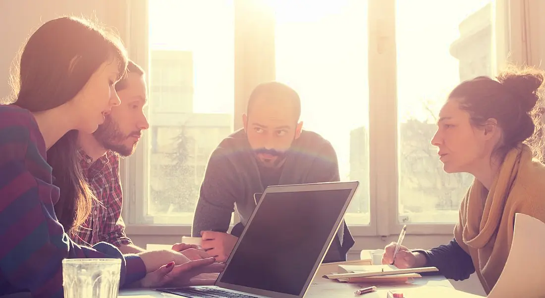 A group of young employees gathered around a table hunched down and talking, trying to avoid using verbal fillers in speech.