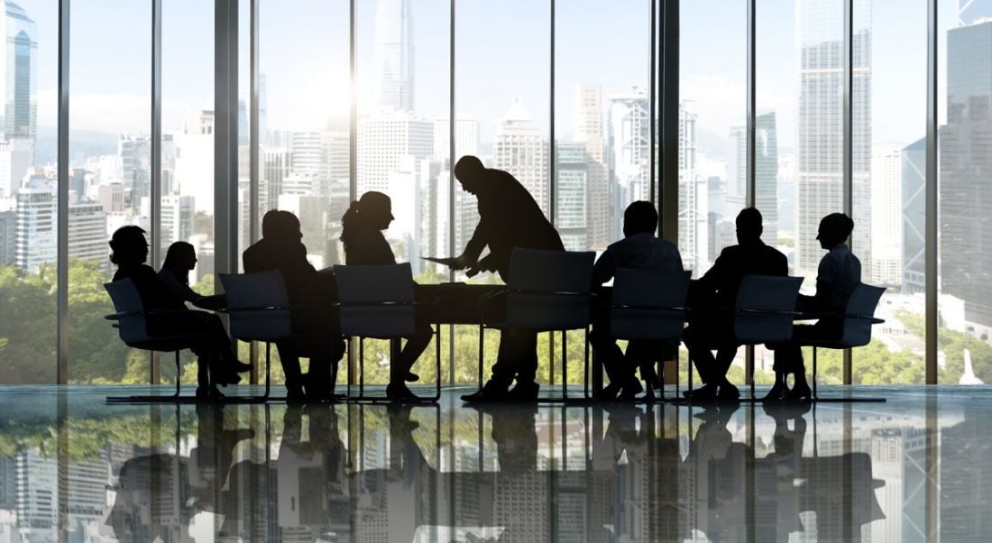 Silhouette of businesspeople crowded around a table in a meeting in a room with ceiling-to-floor windows.