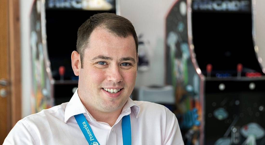 A portrait photo of a brown-haired man with a buzz cut wearing a powder pink shirt and a blue lanyard that says Pramerica on it.