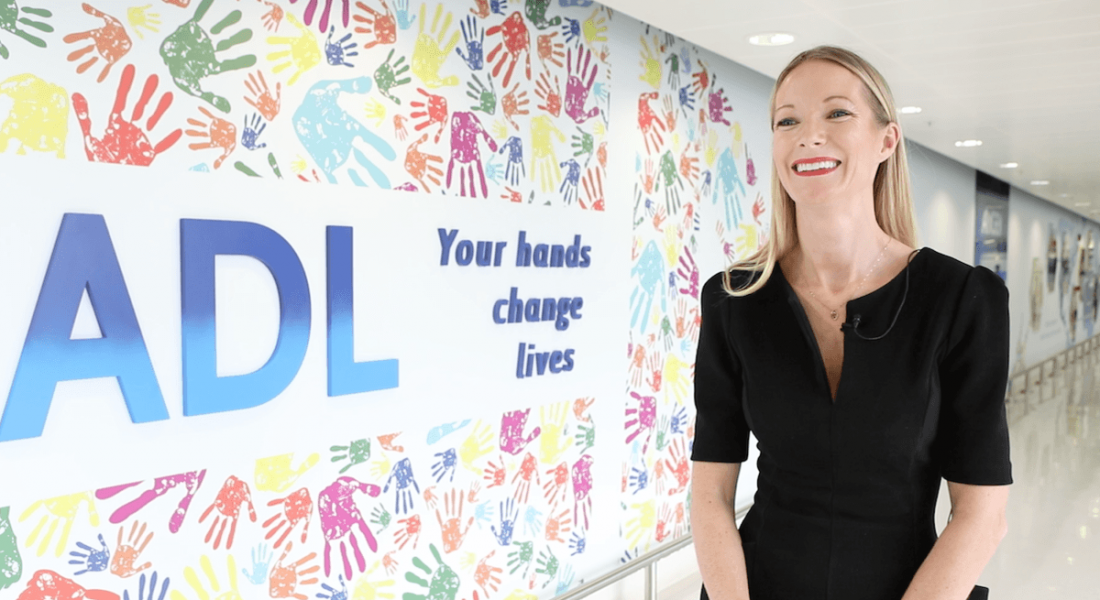 A blonde woman in a black dress smiling, standing beside a large sign with a lot of colourful handprints on it in Amgen.