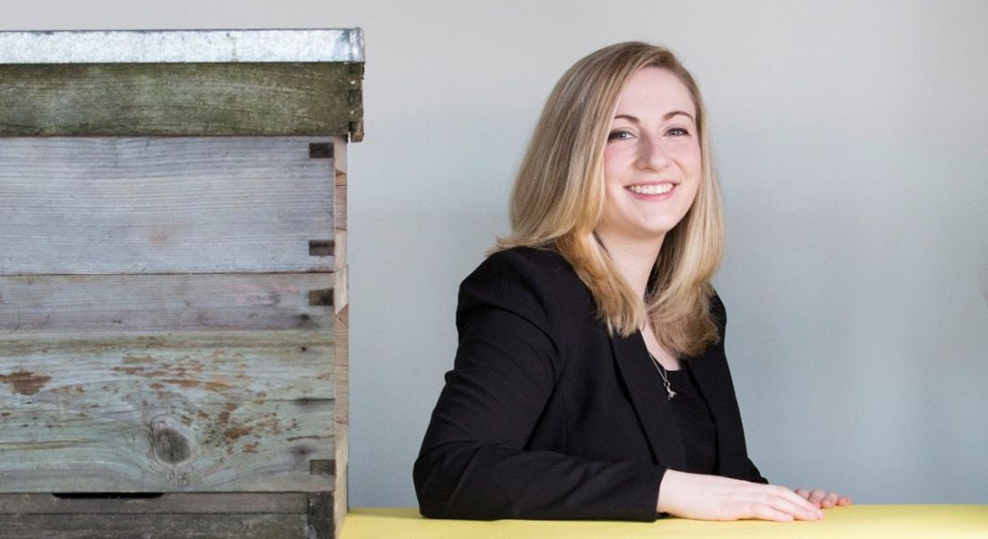 Smiling woman with blonde hair and black jacket beside a bee hive.