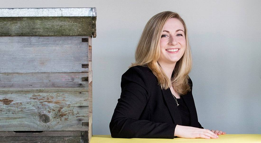 Smiling woman with blonde hair and black jacket beside a bee hive.