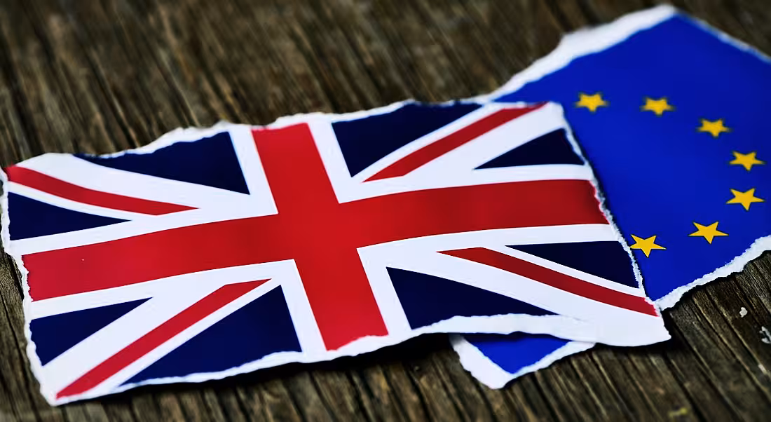 Two torn pieces of paper, one showing the UK flag, the other showing the EU flag on a wooden table, representing Brexit.